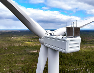 Lähikuvassa tuulivoimalan konehuone ja lavat, taustalla metsää ja taivasta.