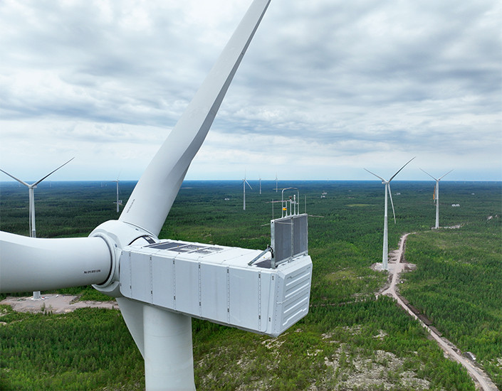 Tuulivoimalan lavat ja konehuone, taustalla metsämaisemaa ja kauempana näkyviä tuulivoimaloita.