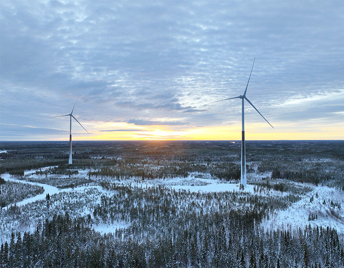 Kaksi tuulivoimalaa talvisessa maisemassa, taustalla laskeva aurinko.