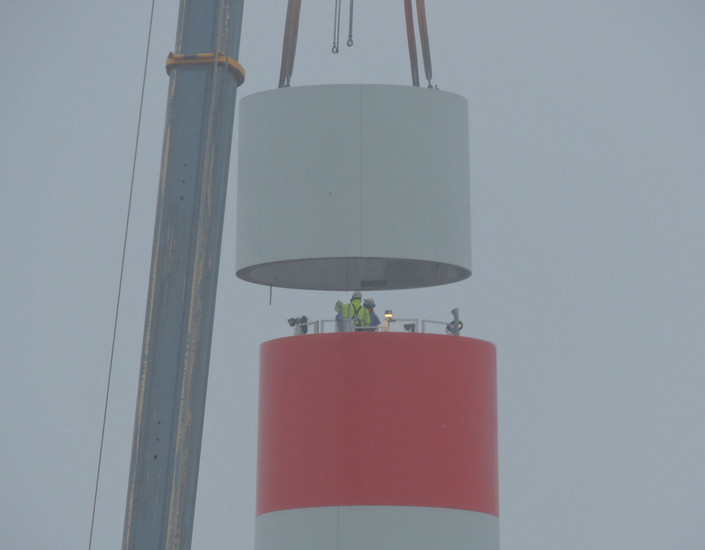 Ein Kran setzt einen Betonring auf den bestehenden Turm, wo er von Arbeitern festmontiert wird