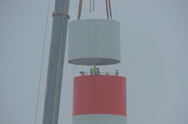 Ein Kran setzt einen Betonring auf den bestehenden Turm, wo er von Arbeitern festmontiert wird